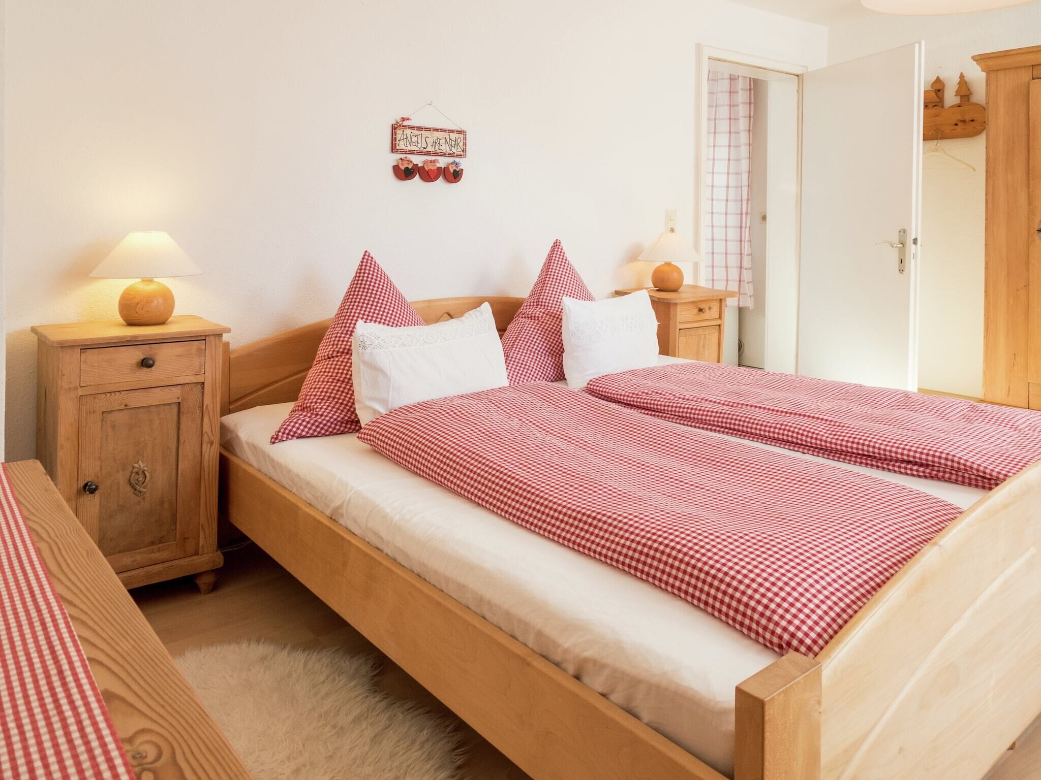 Una cama doble con colcha a cuadros, una mesita de noche de madera con lámpara, un archivador pequeño y un adorno en la pared.