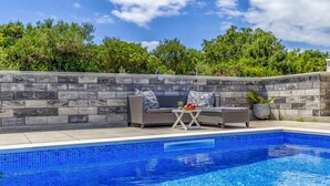 A swimming pool with a stone wall and two wicker sofas with patterned cushions, a small table with fruit, and a potted plant.