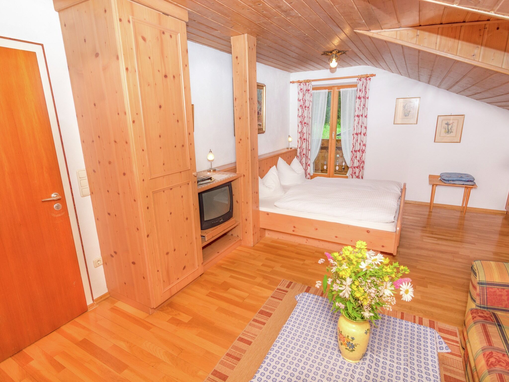 Une chambre avec un plafond en bois, un lit, une télévision, une armoire en bois et une petite table sur laquelle est posé un vase de fleurs.