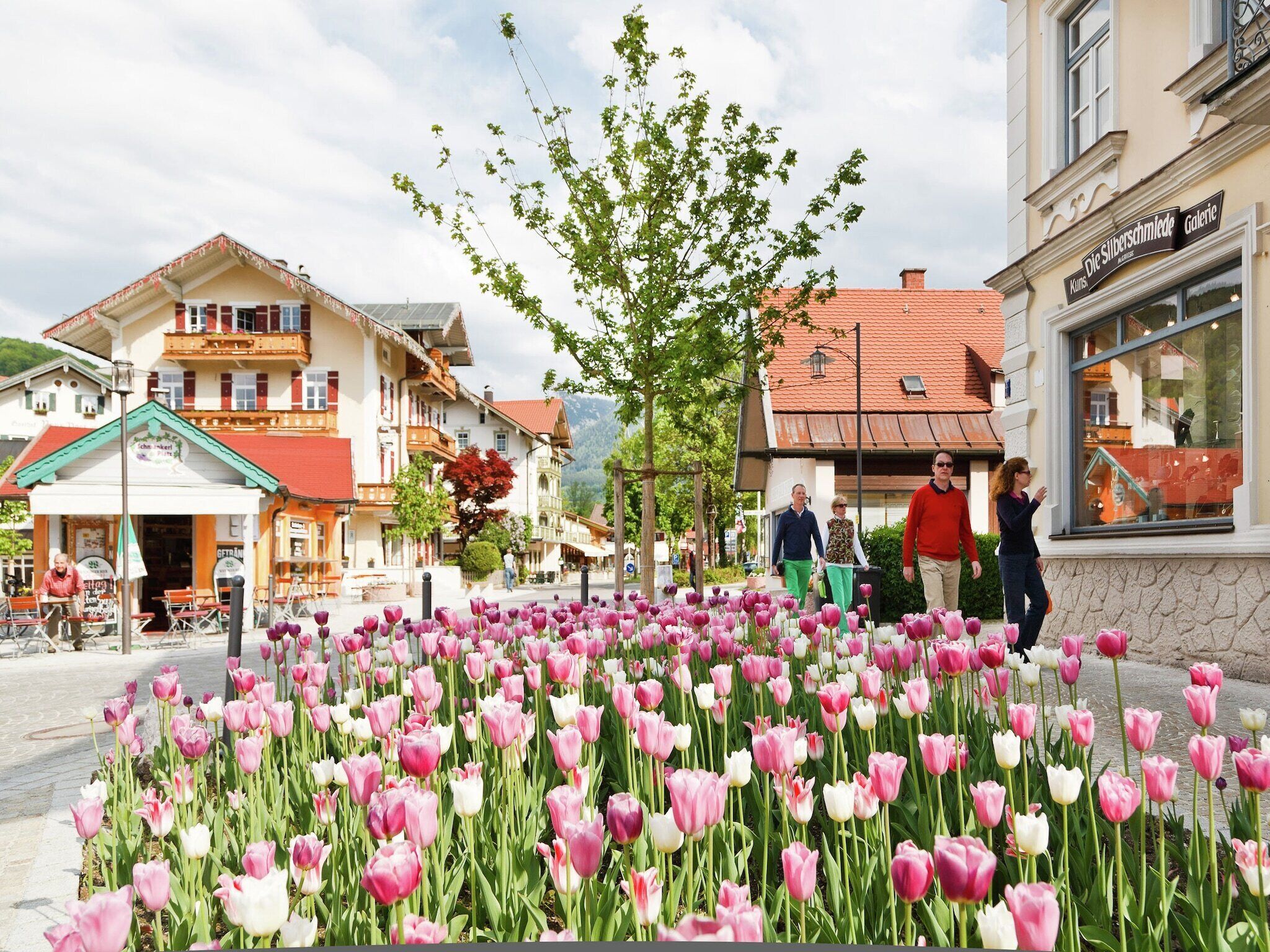Une rue bordée de tulipes roses et blanches au premier plan, des immeubles avec des balcons et des enseignes, et quelques personnes qui se promènent.