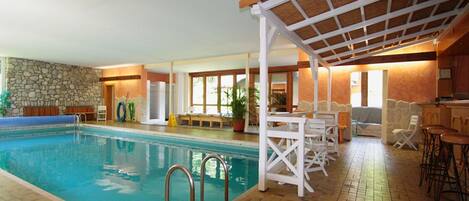 An indoor swimming pool with a stone wall, wooden deck, and a covered seating area with tables and chairs.