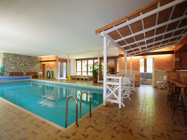 Una piscina interior con pared de piedra, cubierta de madera y zona de estar techada con mesas y sillas.