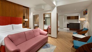 A modern hotel room with a large bed, a pink sofa, a small dining area, and a kitchen in the background. - Ramada Encore By Wyndham Viramgam (Viramgam)