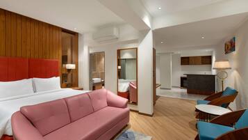 A modern hotel room with a large bed, a pink sofa, a small dining area, and a kitchen in the background.