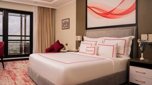 A hotel room with a large bed, a red chair, a nightstand with a lamp, a vase with flowers, and a view through a window.