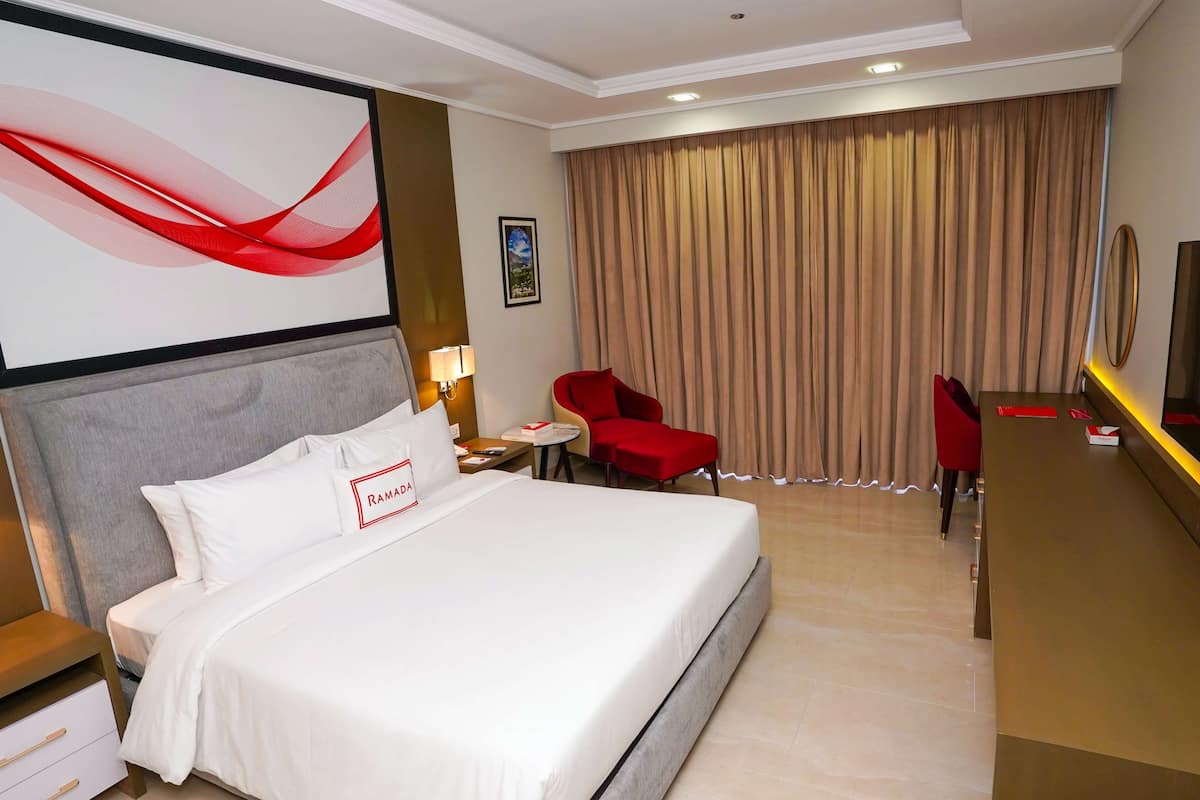 A hotel room with a large bed, a desk, two red chairs, a desk lamp, and a wall-mounted TV.