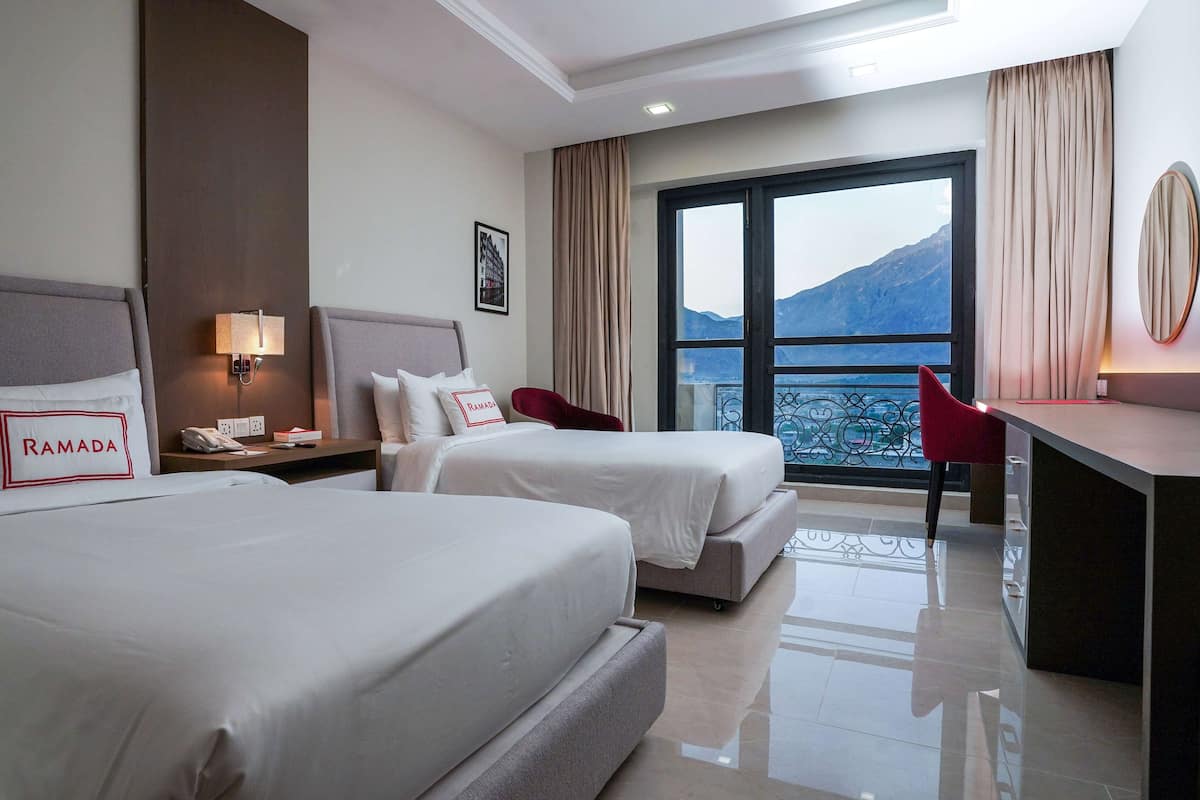 A hotel room with two beds, a desk, a chair, a phone, and a view of mountains through the window.