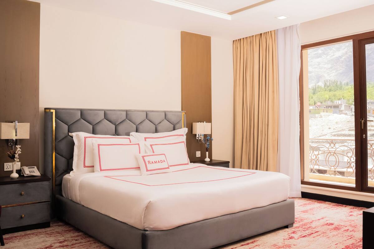 A hotel room with a large bed, a nightstand, a lamp, and a view of a mountainous landscape through the window.