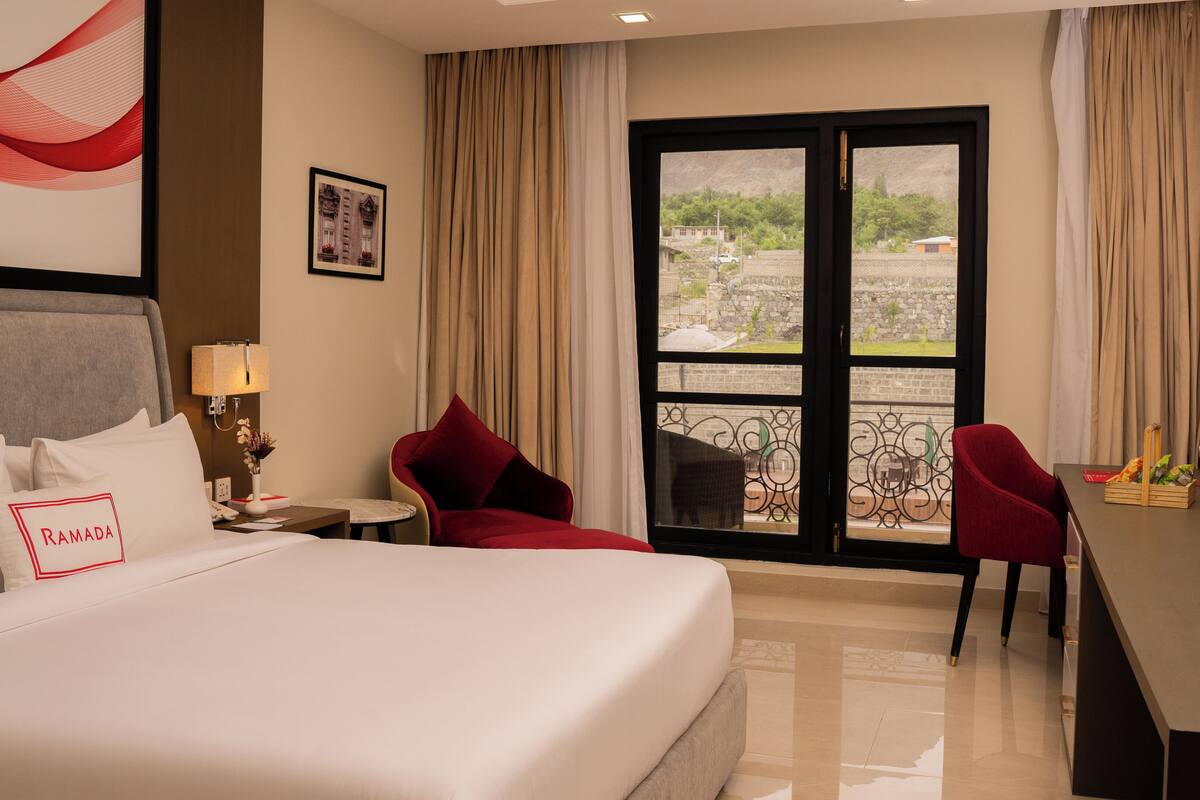 A hotel room with a large bed, a desk, two red chairs, a window with a view of buildings, and a framed picture on the wall.