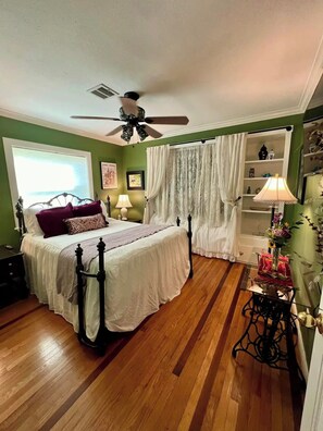 Un dormitorio con cama, ventilador de techo, ventana con persianas, mesita de noche y cómoda.