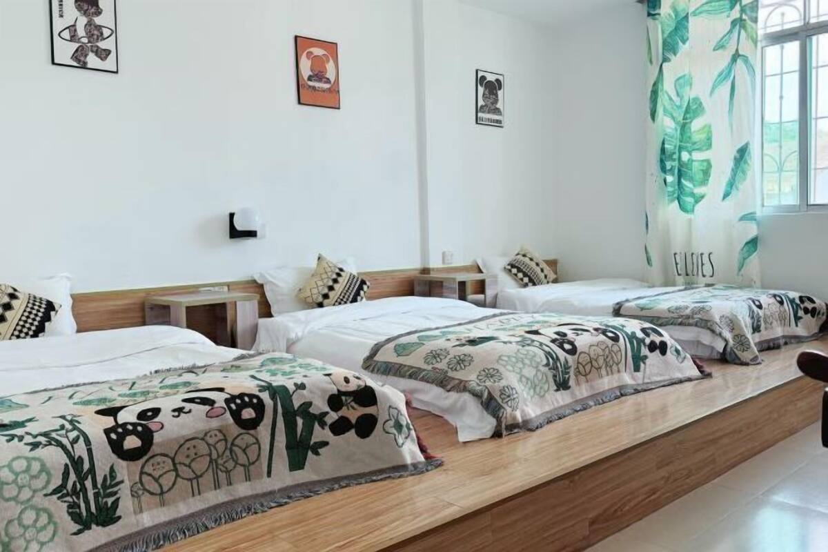 Et hotelværelse med tre senge, hver med mønstret sengetøj, en træbænk og et vindue med gardin.