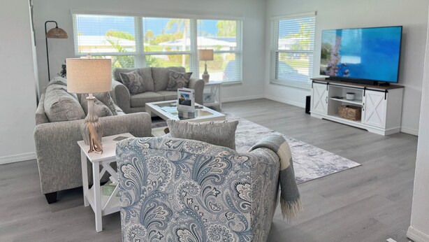 A modern living room with a grey sofa, patterned armchair, and a flat-screen TV on a white cabinet. - Waterfront Retreat 4 BR, 3 BA screened & heated pool, gorgeous canal views (Rotonda West)