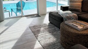 A modern living room with a grey sofa, a wicker ottoman, and a glass door leading to a pool area. - Waterfront Retreat 4 BR, 3 BA screened & heated pool, gorgeous canal views (Rotonda West)