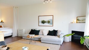 A modern living room with a sofa, a dining table with chairs, a fireplace, and a framed picture on the wall. - Goodview Suite - licensed waterfront studio with a fireplace and patio (Tofino)