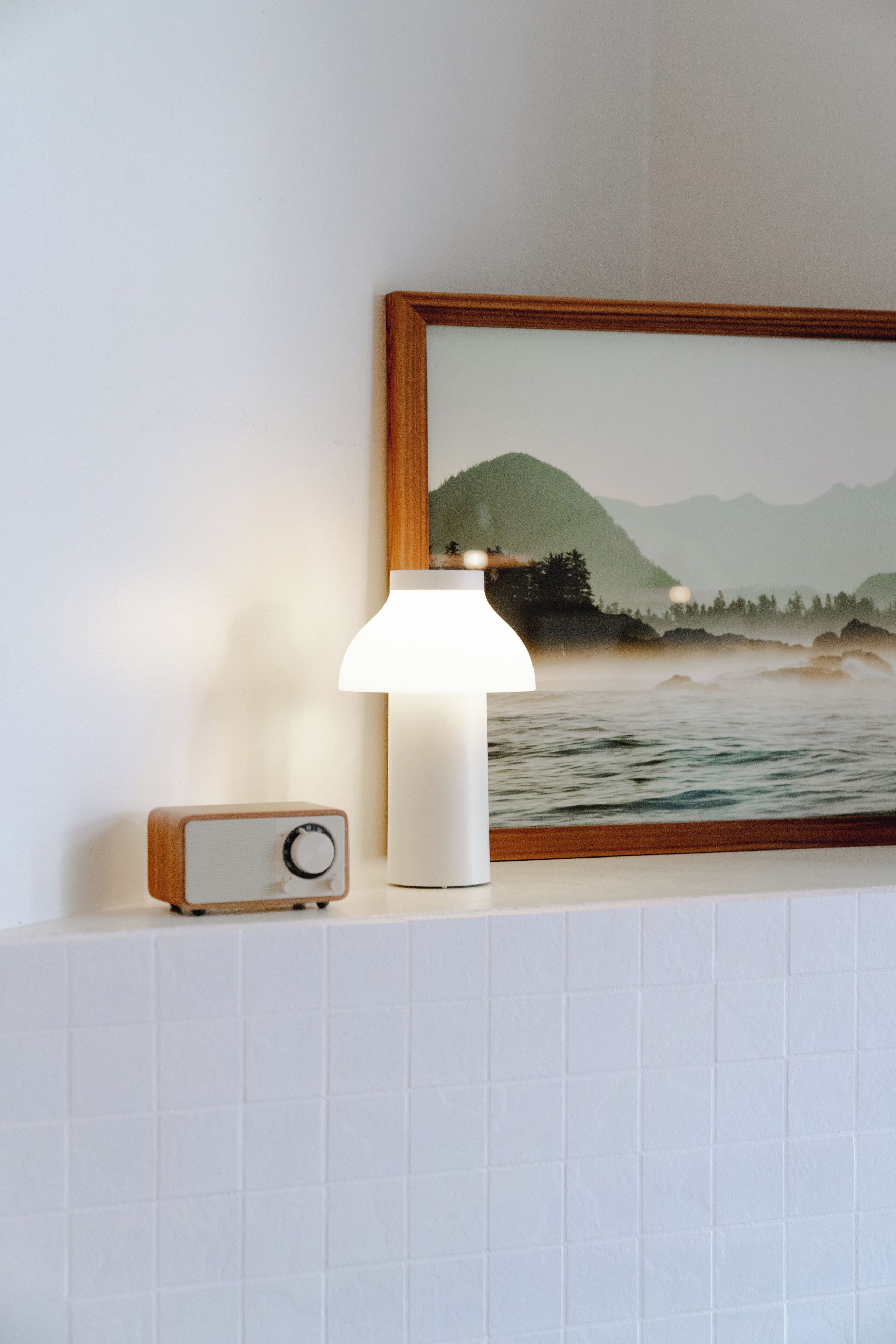 Une radio fixée au mur et une lampe posée sur une surface carrelée, avec un tableau représentant un paysage encadré au-dessus.