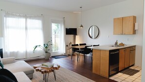 A modern kitchen with wooden cabinets, a dining area with a table and chairs, and a living room with a sofa and a coffee table. - Goodview Suite - licensed waterfront studio with a fireplace and patio (Tofino)