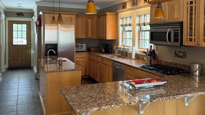 Eine großzügige Küche mit Granitarbeitsplatten, Holzausziehtischen, einer zentralen Kochinsel, Pendelleuchten und einem Edelstahlkühlschrank.