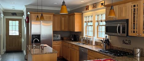 Une cuisine spacieuse dotée de plans de travail en granit, d’armoires en bois, d’un îlot central, de luminaires suspendus et d’un réfrigérateur en acier inoxydable.