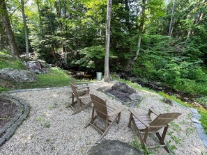 Un área boscosa con un patio de grava, sillas Adirondack de madera y un pequeño estanque.