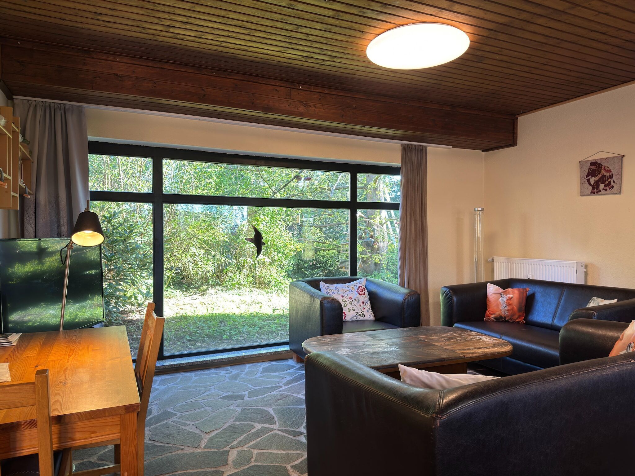 A cozy living room with a stone floor, a wooden table, two leather sofas, a lamp, and a large window with a view of greenery.