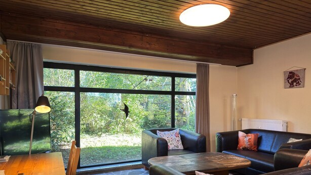 Ett mysigt vardagsrum med ett stengolv, ett träbord, två lädersoffor, en lampa och ett stort fönster med utsikt över grönområden.