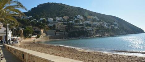 Escena costera con un muro de piedra, playa y edificios en una colina.