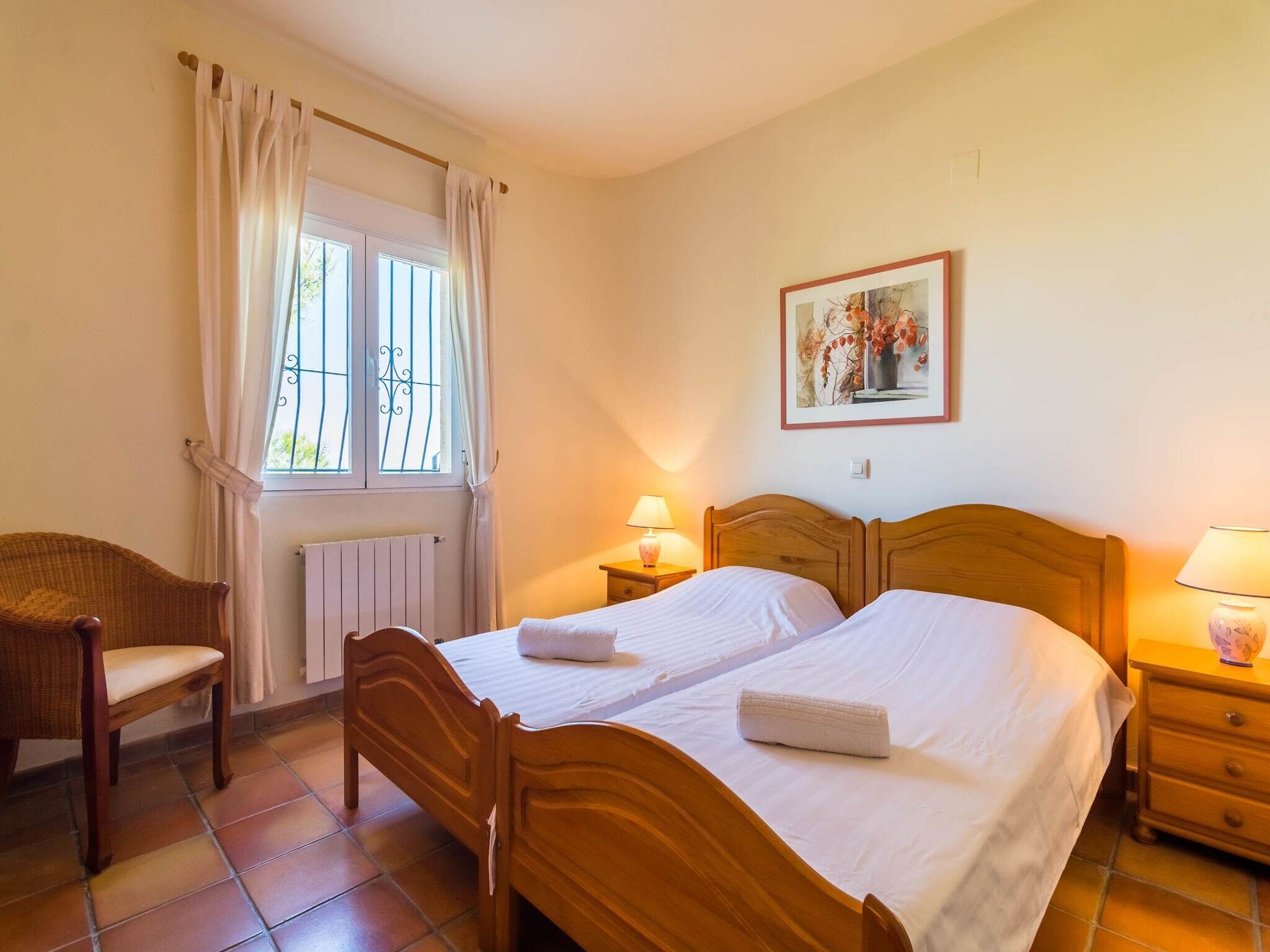 A bedroom with a wooden bed, two bedside tables with lamps, a wicker chair, a window with curtains, and a radiator.