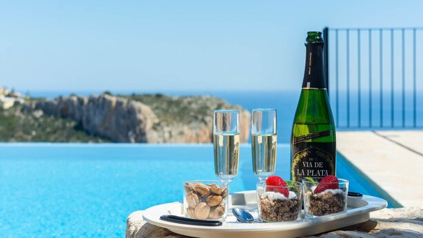Une bouteille de champagne, deux verres et un plateau avec des noix et des céréales sur un rebord en pierre, près d’une piscine offrant une vue sur des falaises.