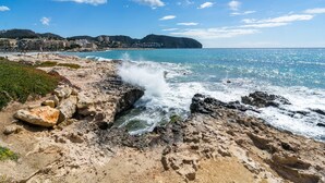 Une côte rocheuse où les vagues s’écrasent sur le rivage, un ciel bleu dégagé et des bâtiments visibles à l’arrière-plan.