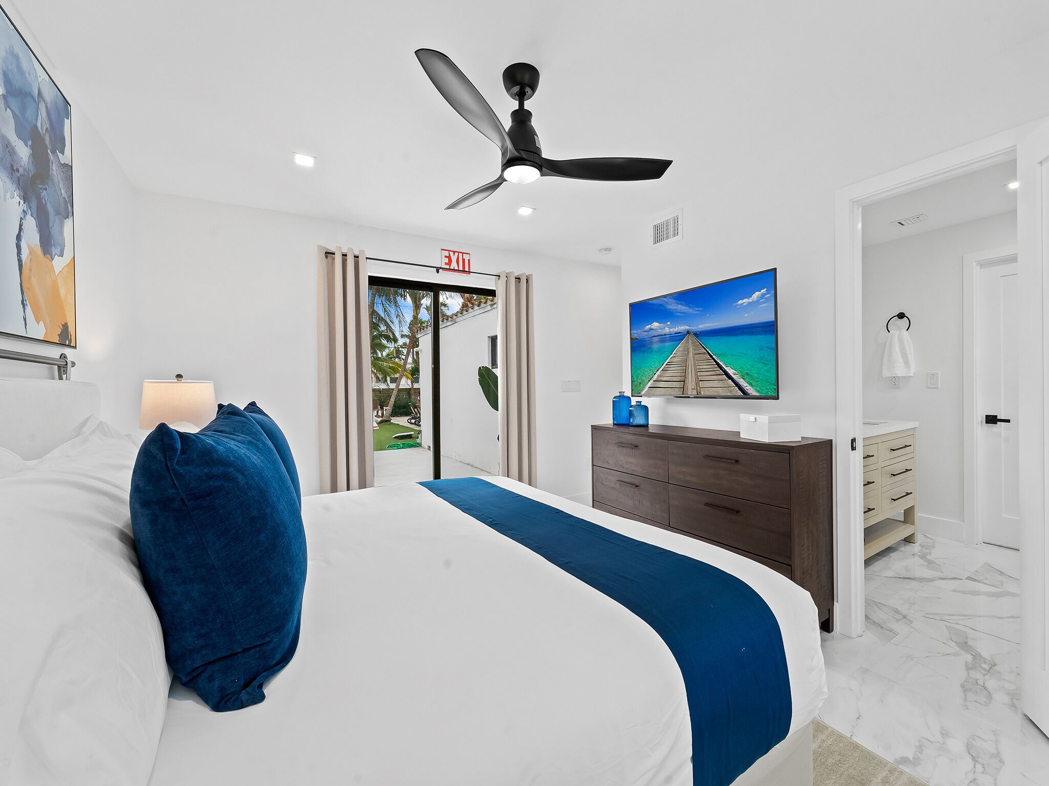 A modern hotel room with a bed, a ceiling fan, a TV, and a view of the ocean through a door.