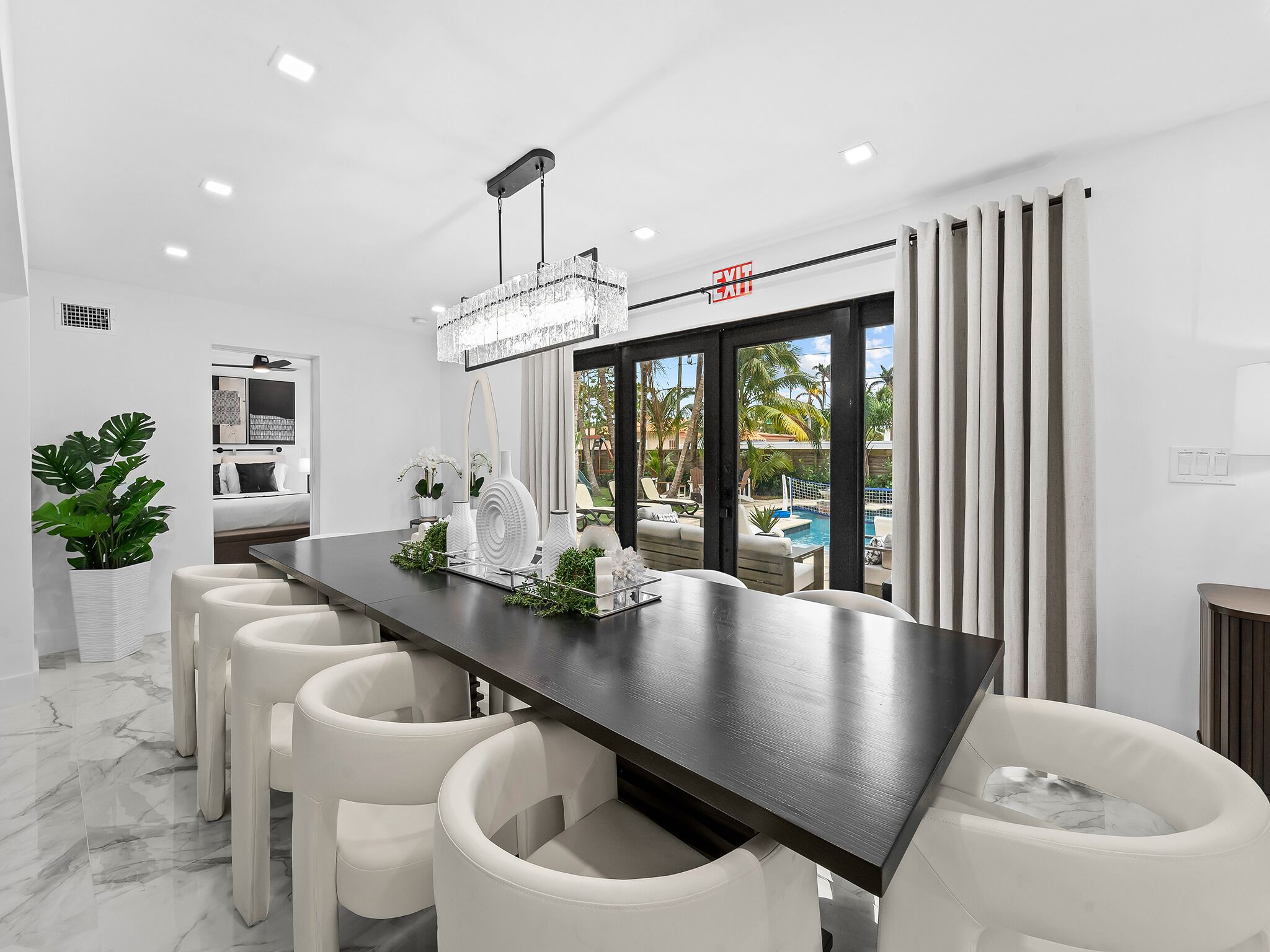 A modern dining area with a marble floor, a long table with white chairs, a chandelier, and a view of a pool and outdoor seating.