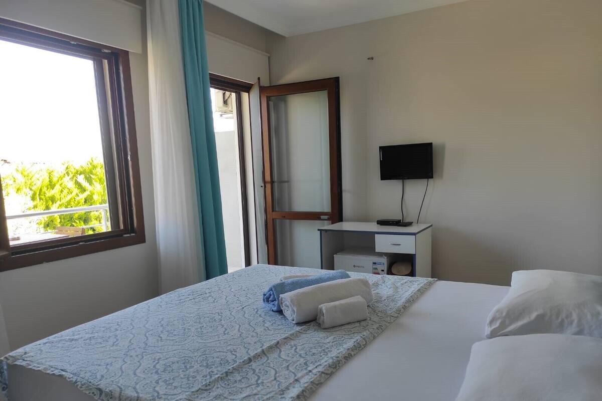 A hotel room with a bed, a desk with a flat-screen TV, a window with curtains, and a view of greenery outside.
