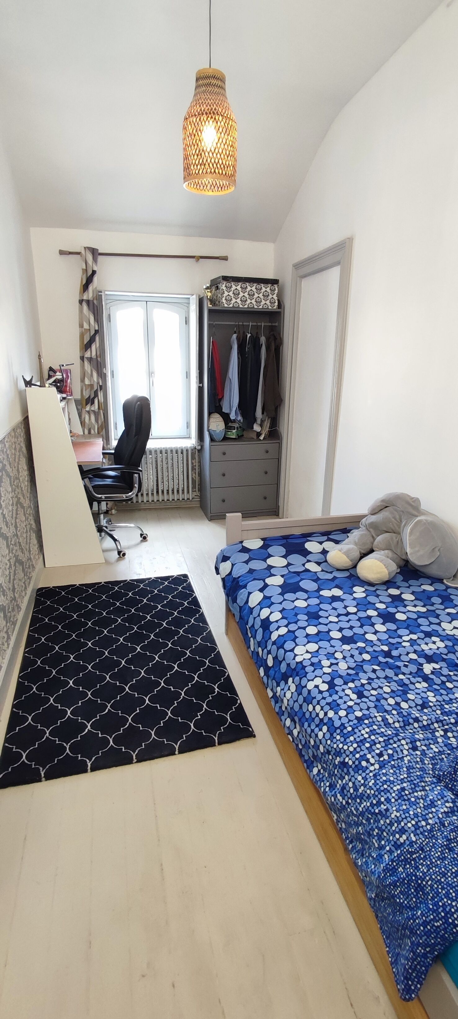 A bedroom with a bed, a desk, a chair, a wardrobe, and a hanging light fixture.