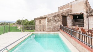 Ein Schwimmbecken mit Fliesen umgeben, ein Gebäude aus Stein mit einem Balkon und Gartenmöbel.
