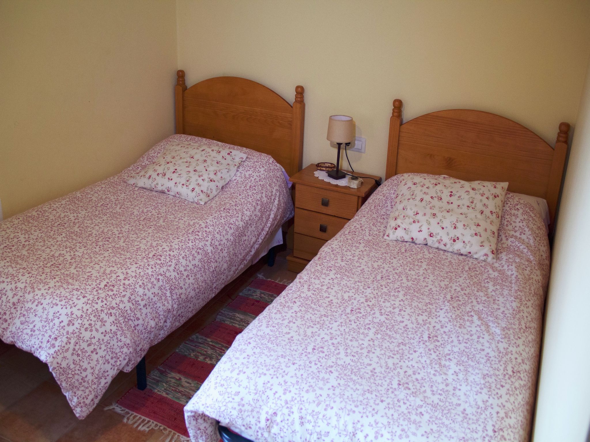 Habitación con dos camas individuales, cada una con cabecero de madera y ropa de cama estampada con motivos florales. Hay una mesita de noche con lámpara entre las camas.