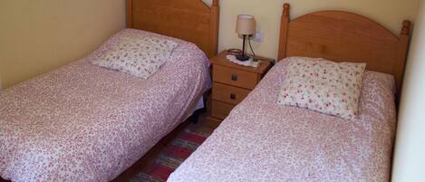 Habitación con dos camas individuales, cada una con cabecero de madera y ropa de cama estampada con motivos florales. Hay una mesita de noche con lámpara entre las camas.