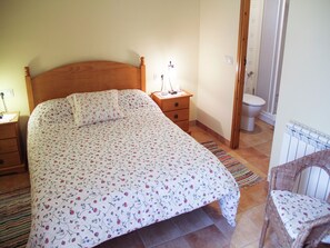 Ein Schlafzimmer mit einem hölzernen Bett, einem Nachttisch, einem Beistelltisch, einem Sessel und einem durchgehenden Türspalte sichtbaren Badezimmer.