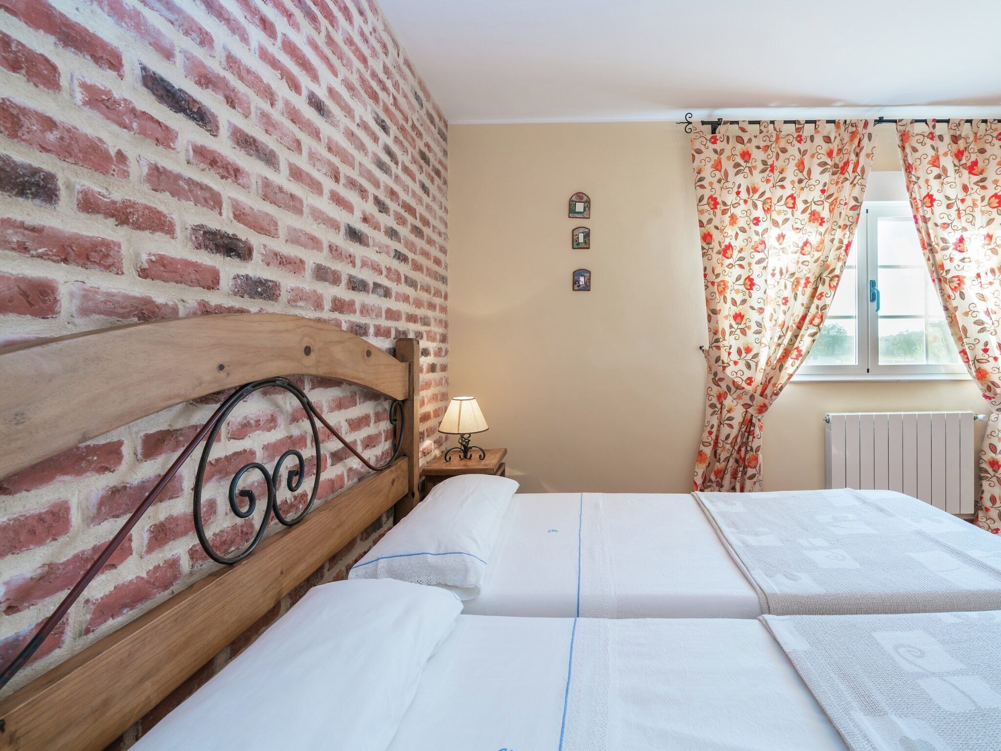 A room with a brick wall, a wooden headboard, a bed with white linens, a small bedside table with a lamp, and floral curtains.
