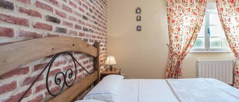 A room with a brick wall, a wooden headboard, a bed with white linens, a small bedside table with a lamp, and floral curtains.