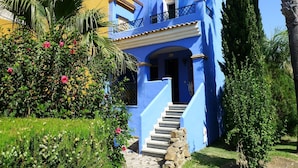 Un immeuble de deux étages avec une façade bleue, un balcon et un escalier en pierre menant à l’entrée.