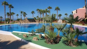 Une piscine entourée de palmiers et bordée d’un mur de pierre, avec un ciel bleu clair en arrière-plan.