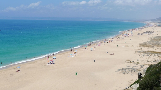 Une vaste plage avec une eau d’un bleu limpide, des parasols et des gens.