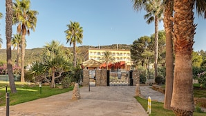 Un bâtiment doté d’une porte en pierre, bordé de palmiers, avec un paysage vallonné en arrière-plan.