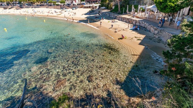 Una playa con agua cristalina, un muro de piedra y edificios al fondo.