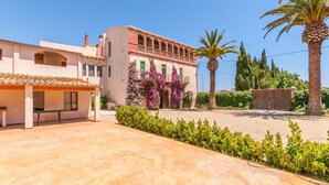 Una villa de estilo mediterráneo con un patio amplio, un porche cubierto y un jardín exuberante con palmeras y plantas en flor.