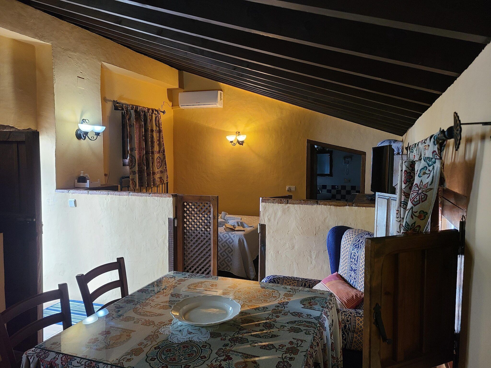 A dining area with a table, chairs, a wall-mounted air conditioner, and a patterned tablecloth.