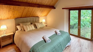 Ein Schlafzimmer mit Holzbalkendecke, einem großen Bett mit Kissen, einem Nachttisch mit Lampe und Blick auf Grün durch ein Fenster.