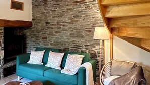 Ein gemütliches Zimmer mit Steinwand, einem grünen Sofa mit gemusterten Kissen, einem Holztisch, einem Korbstuhl und einem Kamin.