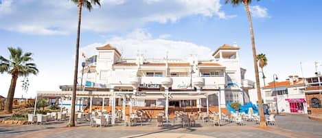 A coastal building with a cafe area in front, palm trees, and a clear sky.
