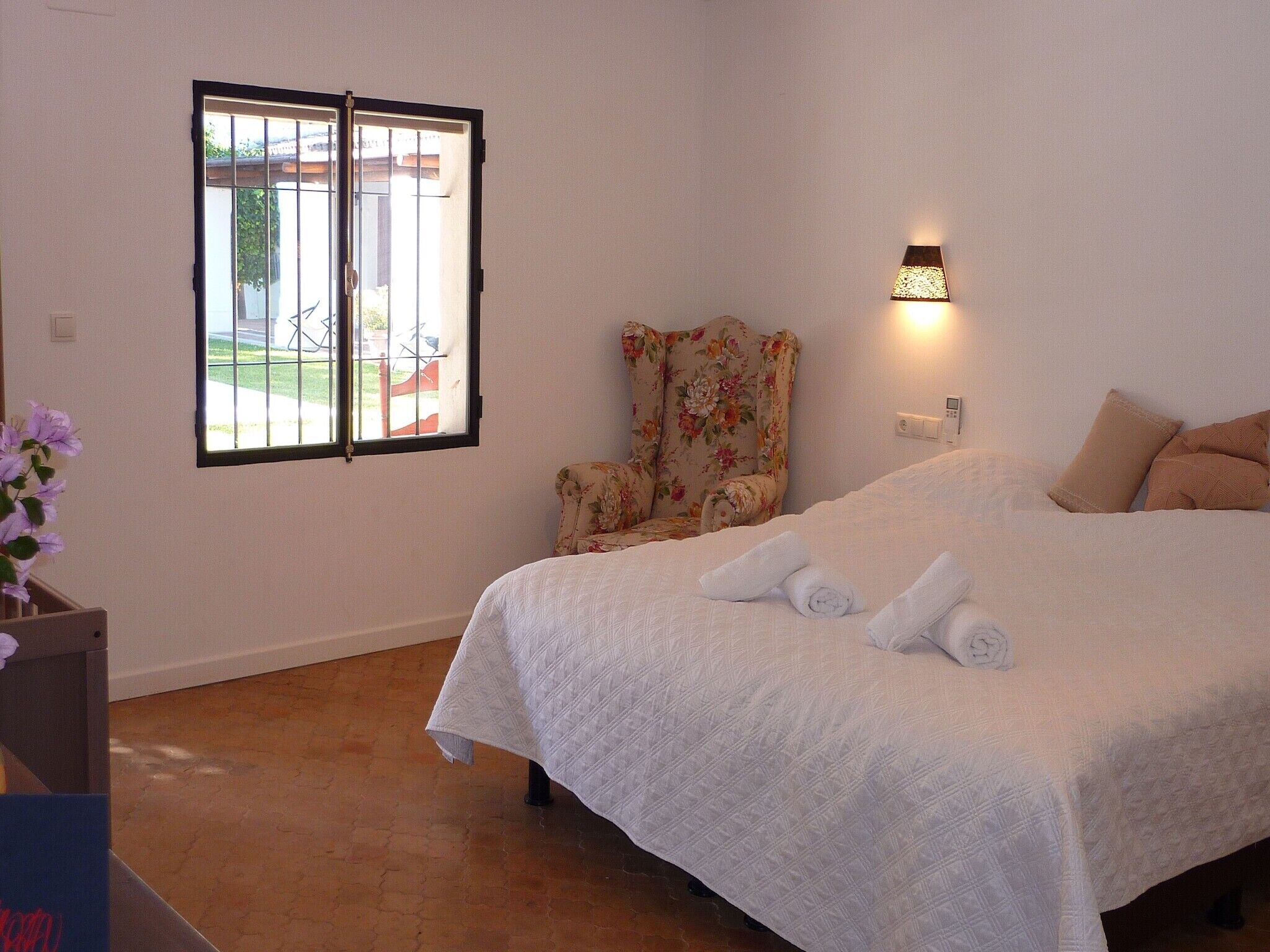 A bedroom with a bed, a chair, a window, and a wall-mounted light.